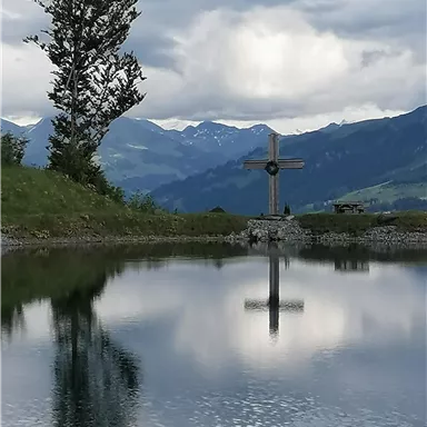 A quiet mountain lake with a cross sculpture in the foreground. The mountains are visible in the background, and the clouds are reflecting in the water.