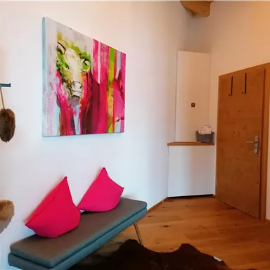A modern hallway with a colorful painting on the wall and a gray bench. Red cushions and a wooden shelf give the room an inviting atmosphere.