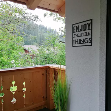 A cozy balcony with wooden paneling and a beautiful view of nature. On the wall hangs a sign that reads "ENJOY THE LITTLE THINGS".
