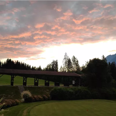 Ein malerischer Sonnenuntergang über den Bergen, mit einem klaren Himmel und sanften Wolken. Im Vordergrund befindet sich eine grüne Wiese und eine rustikale Holzkonstruktion.