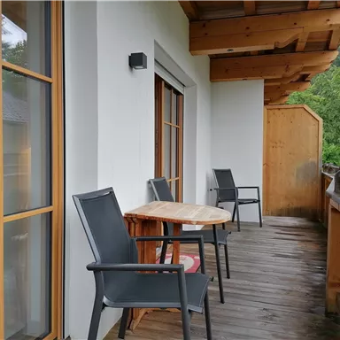 Ein gemütlicher Balkon mit einem Holztisch und zwei Stühlen. Im Hintergrund sind grüne Bäume sichtbar.