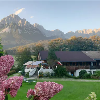 Eine malerische Landschaft mit Bergen im Hintergrund und einem gemütlichen Gebäude. Im Vordergrund blühen pinkfarbene Blumen auf einer grünen Wiese.