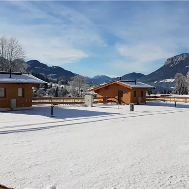 Winterlandschaft mit Holzhütten und schneebedecktem Boden. Im Hintergrund sind Berge und ein klarer Himmel zu sehen.