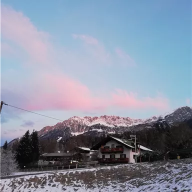 A winter landscape with snow-covered mountains and a pretty house. The sky displays gentle shades of pink and blue.