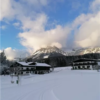 A winter landscape with snow-covered hills and a clear blue sky. In the foreground, some traditional buildings can be seen.