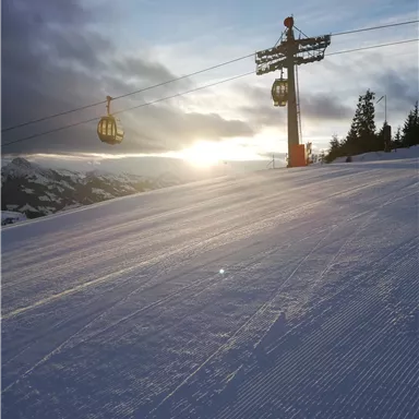A ski landscape with freshly groomed slopes and a cable car support structure. The sunset illuminates the scene in warm colors.