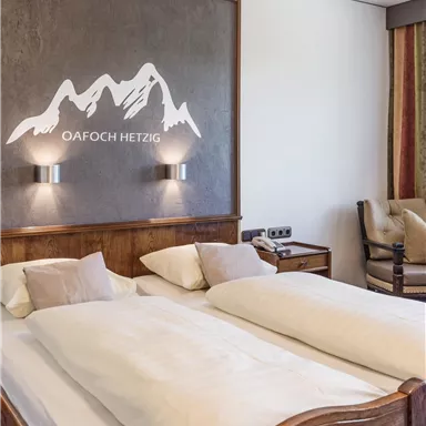 A cozy bedroom with two beds and beautiful wooden furniture. On the wall is a mountain silhouette with the inscription "Oafach Hetzig."