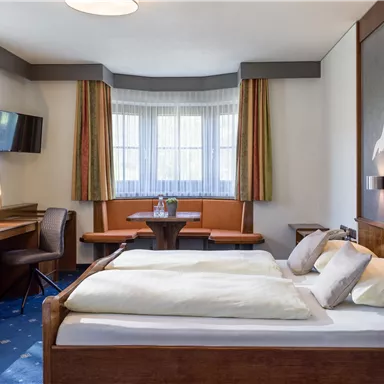 A cozy room with a large bed, a table, and chairs. Bright windows provide natural light and a pleasant atmosphere.