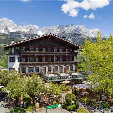 A charming hotel with traditional architecture, surrounded by green trees. Majestic mountains rise in the background beneath a clear sky.