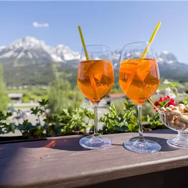 Two glasses with an orange drink are on a table overlooking snow-covered mountains. Next to the glasses is a bowl of snacks.