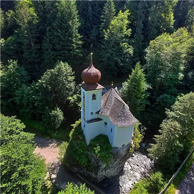 A picturesque chapel on a small rocky outcrop, surrounded by dense forests. The dome has a striking red color and the building exudes tranquility.
