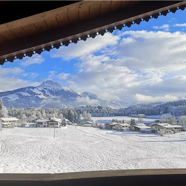 Eine winterliche Landschaft mit schneebedeckten Feldern und Hügeln. Im Hintergrund sind Berge und ein blauer Himmel mit einigen Wolken zu sehen.