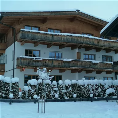 Ein gemütliches Chalet im Schnee mit Holzbalkonen. Die Umgebung ist von schneebedeckten Bäumen und Sträuchern umgeben.
