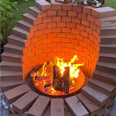 A masonry fire pit with a roaring fire and logs. In the background, green plants are visible.