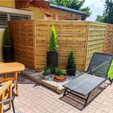 A cozy terrace area with a table, chairs, and a lounger. Surrounded by green plants and a wooden privacy screen.
