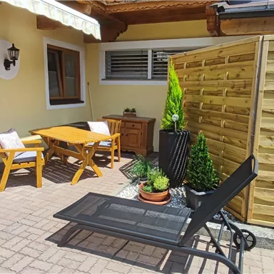 A cozy terrace with a wooden table and chairs. In the background, there is a wooden carving wall and some plants.
