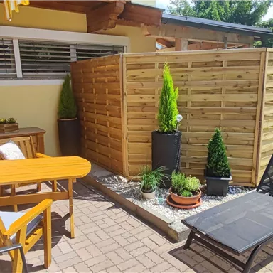 A cozy outdoor area with a table, chairs, and a lounge chair. Surrounded by plants and a wooden privacy fence.