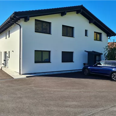 Ein modernes, zweigeschossiges Haus mit einer hellen Fassade steht in einer ruhigen Umgebung. Ein Auto parkt auf dem gepflasterten Weg vor dem Gebäude.