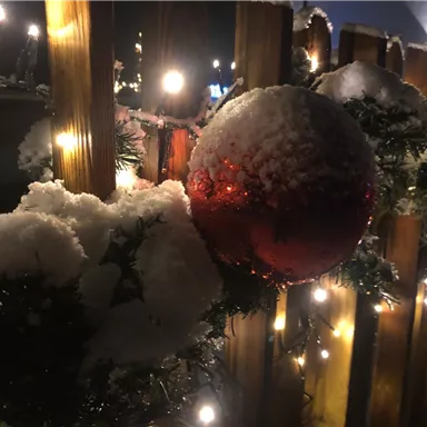 A shiny red Christmas ornament hangs on a wintry fence adorned with snow and pine branches. In the background, warm lights glow in the darkness.
