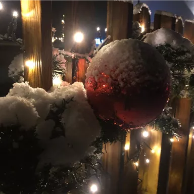 Eine rot glänzende Christbaumkugel hängt an einem winterlichen Zaun, der mit Schnee und Tannenzweigen geschmückt ist. Im Hintergrund leuchten warme Lichter in der Dunkelheit.
