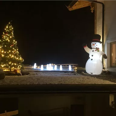 A festively decorated terrace at night with a glowing Christmas tree and a plastic snowman. The ground is covered in snow, and some lights shine in the background.