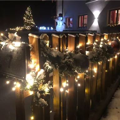 A festively decorated fence with lights and Christmas decorations in the snow. In the background, there is a snowman and a Christmas tree is glowing.