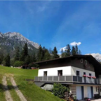 Ein modernes Haus in einem malerischen Tal, umgeben von hohen Bergen und grünen Wiesen. Der Himmel ist klar und blau, was eine ruhige Atmosphäre schafft.