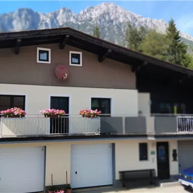 Ein modernes Haus mit Balkon und Blumenkästen inmitten einer malerischen Landschaft. Im Hintergrund sind grüne Wiesen und hohe Berge zu sehen.