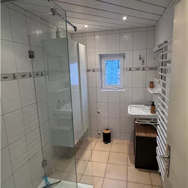 A modern bathroom with a glass shower and a sink. Bright tiles and a large window create an airy atmosphere.