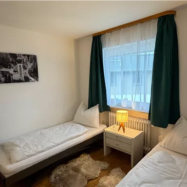 A simple room with two single beds and a nightstand. The curtains are green and there is a warm light from a lamp.