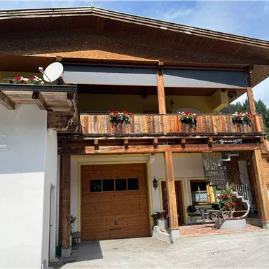 A traditional wooden house with a balcony and blooming plants. The facade is bright and welcoming, surrounded by a picturesque landscape.