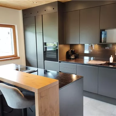 A modern kitchen with dark cabinets and an elegant countertop. At the table, there are chairs, and daylight is coming in through the window.