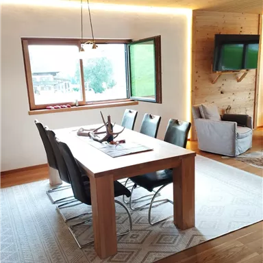 A modern dining room with a wooden table and black chairs. Large windows offer a view of the landscape.