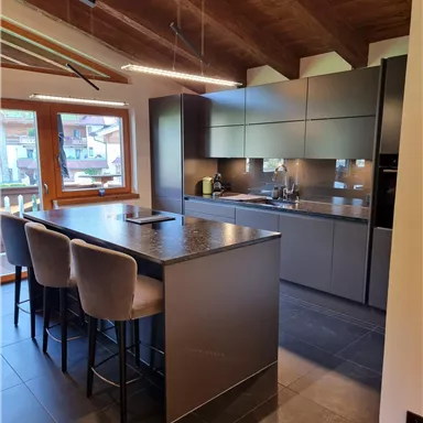 Modern kitchen with dark furniture and a large kitchen island. Large windows let in plenty of daylight and offer a view outside.