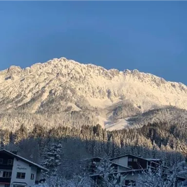 A snow-covered mountain landscape under a clear blue sky. In the foreground, some houses and trees are visible.