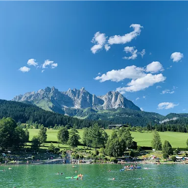 Ein klarer See umgeben von grünen Wiesen und hohen Bergen. Der Himmel ist blau mit einigen Wolken.