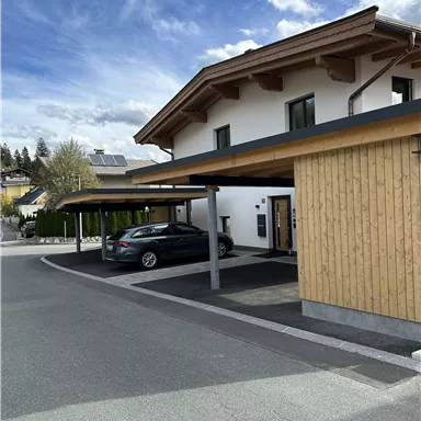 A modern house with wooden cladding and a covered parking space. The sky is blue with some clouds.