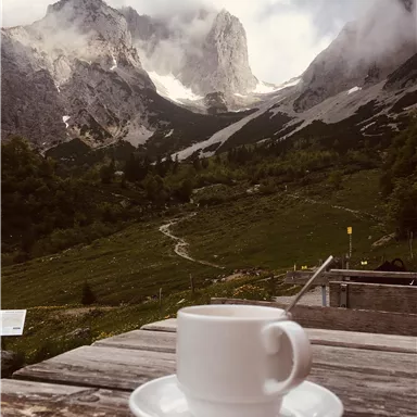 Eine Tasse Kaffee auf einem Holztisch mit einer majestätischen Berglandschaft im Hintergrund. Nebel umhüllt die Gipfel und die Natur ist wunderschön grün.
