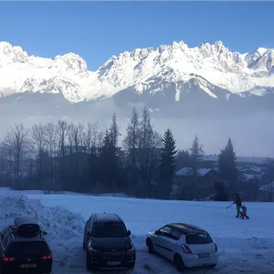 A snow-covered landscape with majestic mountains in the background. In the foreground, there are several cars and a person walking in the snow.