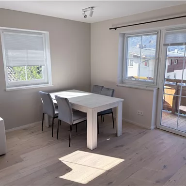 A bright, modern dining area with a white table and gray chairs. Large windows provide a view outside and let in plenty of daylight.