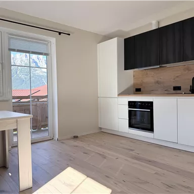 A modern kitchen unit with white cabinets and a wooden workspace. In the background, there are windows with a mountain view and a light wooden floor.