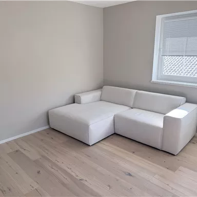 A modern, bright living room with a white sofa and a window. The floor is made of light wood, and the walls are painted in a neutral shade.