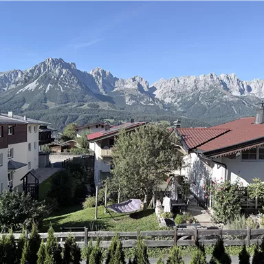 A picturesque residential area with houses and a garden. In the background, majestic mountains rise under a clear blue sky.