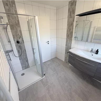 A modern bathroom with shower and sink. The walls are light tiled and there is a towel radiator.