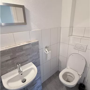 A modern bathroom with a white toilet and a small sink. The walls are lined with light tiles.