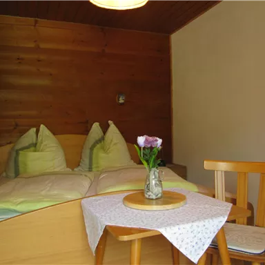 A cozy bedroom with a double bed and two pillows. A small table with a vase and flowers complements the decor.