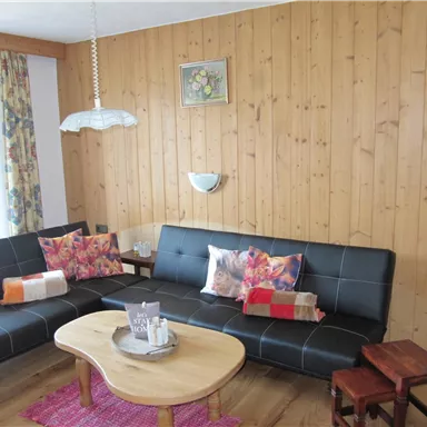 A cozy living room with wooden paneling and two black sofas. In the center is a round table with decorative elements and colorful cushions on the sofas.
