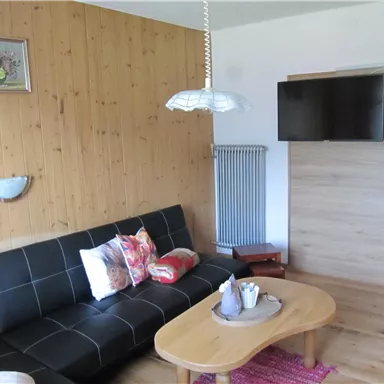 A cozy living room with a black sofa and colorful pillows. A television hangs on the wall and next to the sofa is a round table.