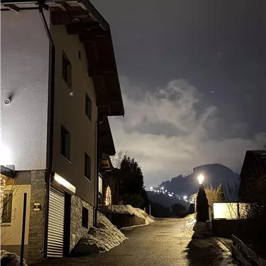 A quiet street at night with a modern building on the left side. In the background, trees and gentle hills are visible, illuminated by a clear sky.