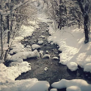 Ein schmaler Bach fließt durch eine schneebedeckte Landschaft. Die Ufer sind mit frischem Schnee und runden Steinen bedeckt.